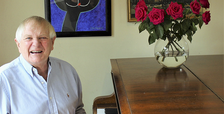 Composer Paul Williams seated at his grand piano on which is a vase of red roses